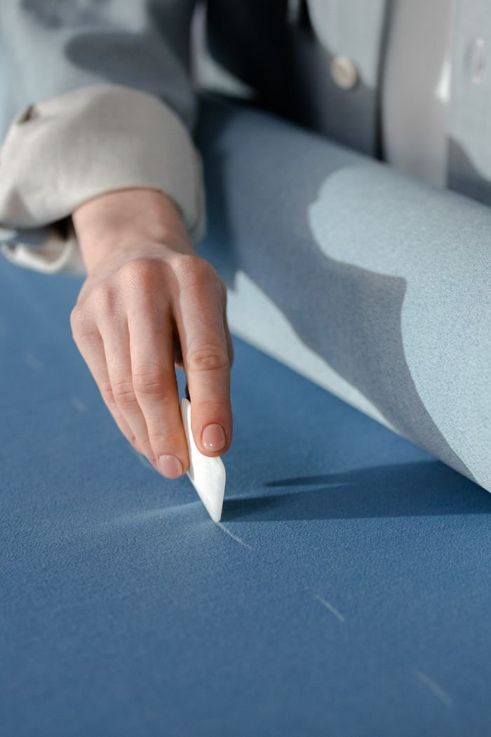 gallery-1 A tailors hand marking blue fabric with chalk, illustrating precision in tailoring.